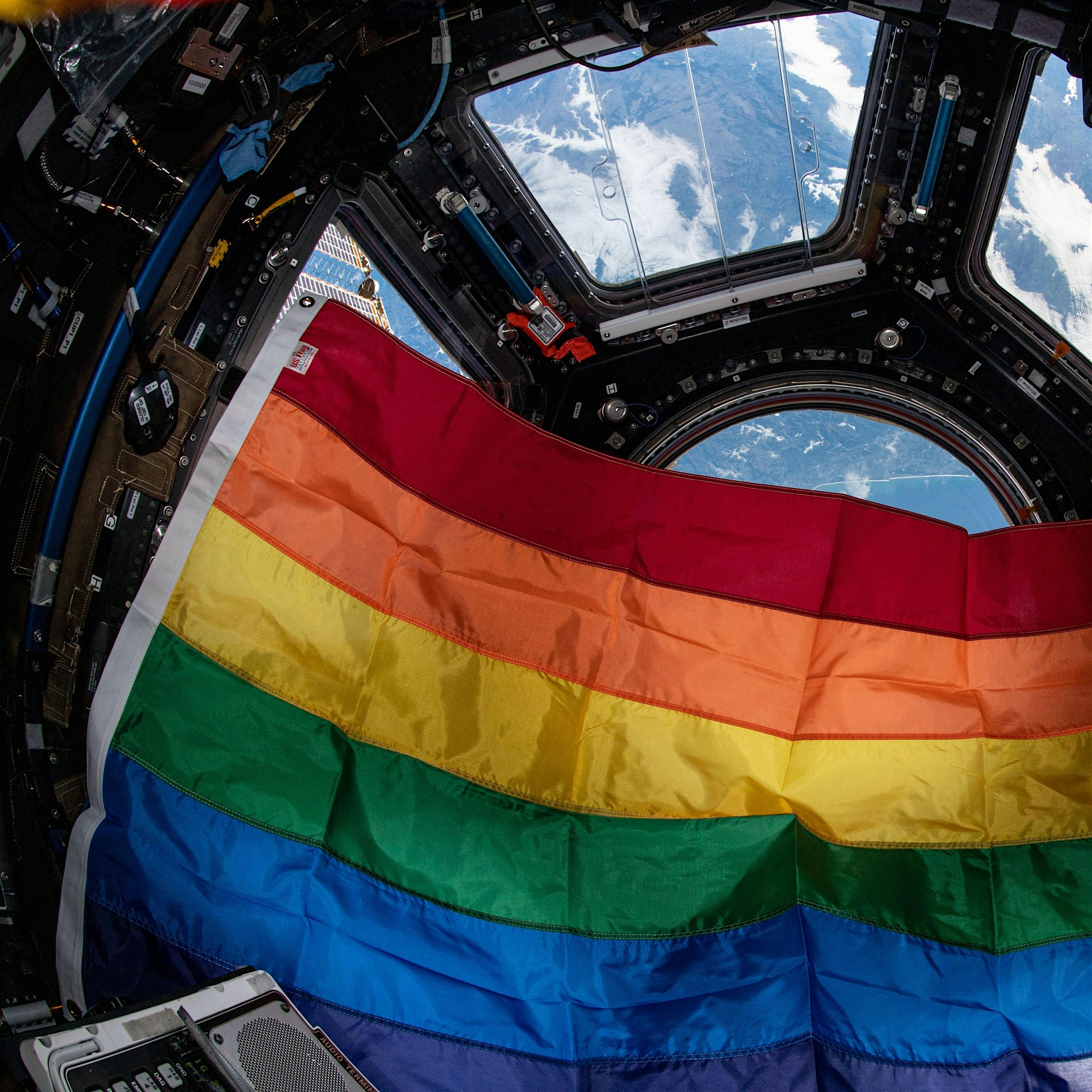 Rainbow Pride flag draped across equipment inside a spacecraft cabin, with Earth visible through a circular window nearby