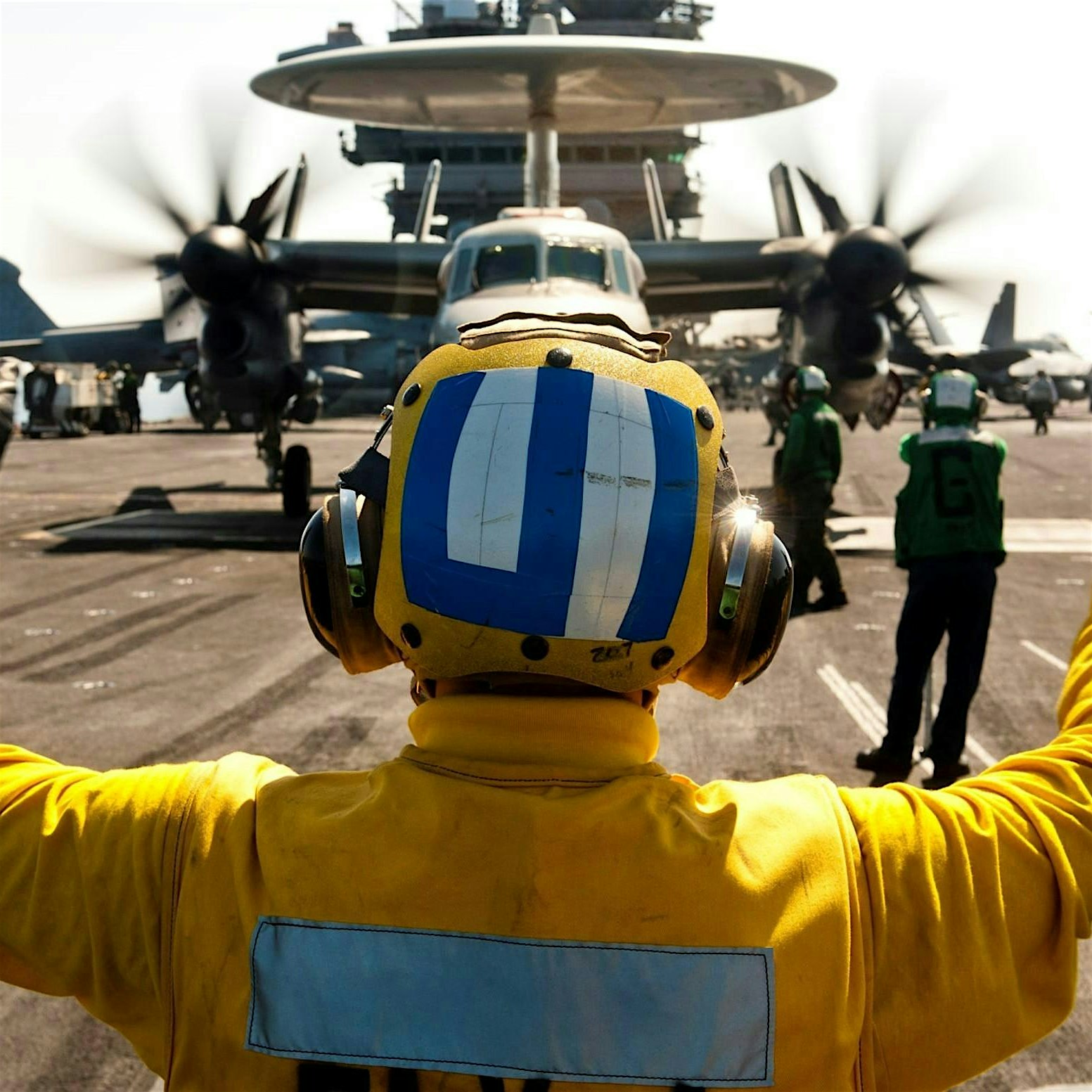 Back view of a yellow flight-suit deck crew member signaling with arms raised on an aircraft carrier, with a large propeller aircraft in the background.