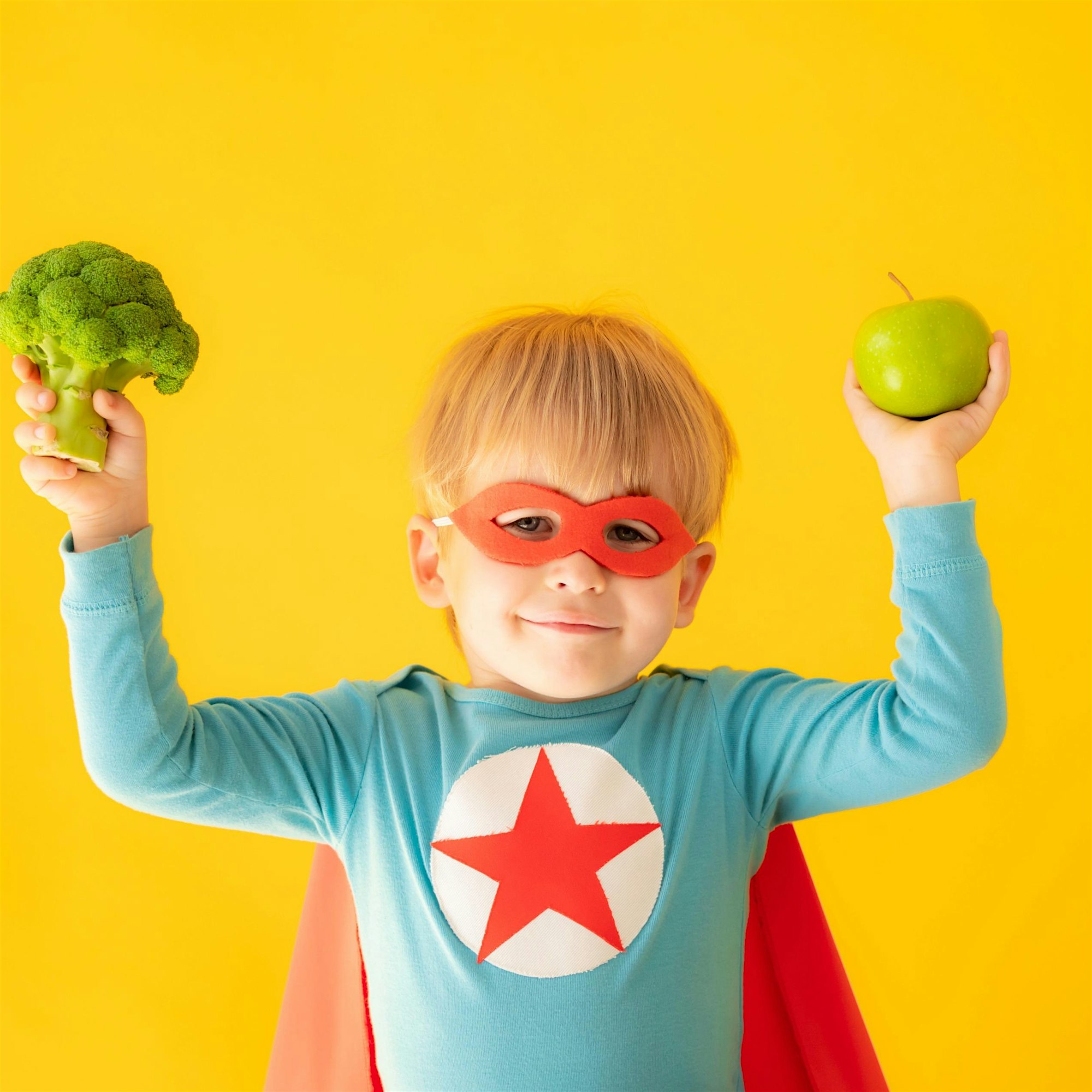 Child wearing a red superhero mask holds broccoli in the left hand and a green apple in the right, against a bright yellow background.