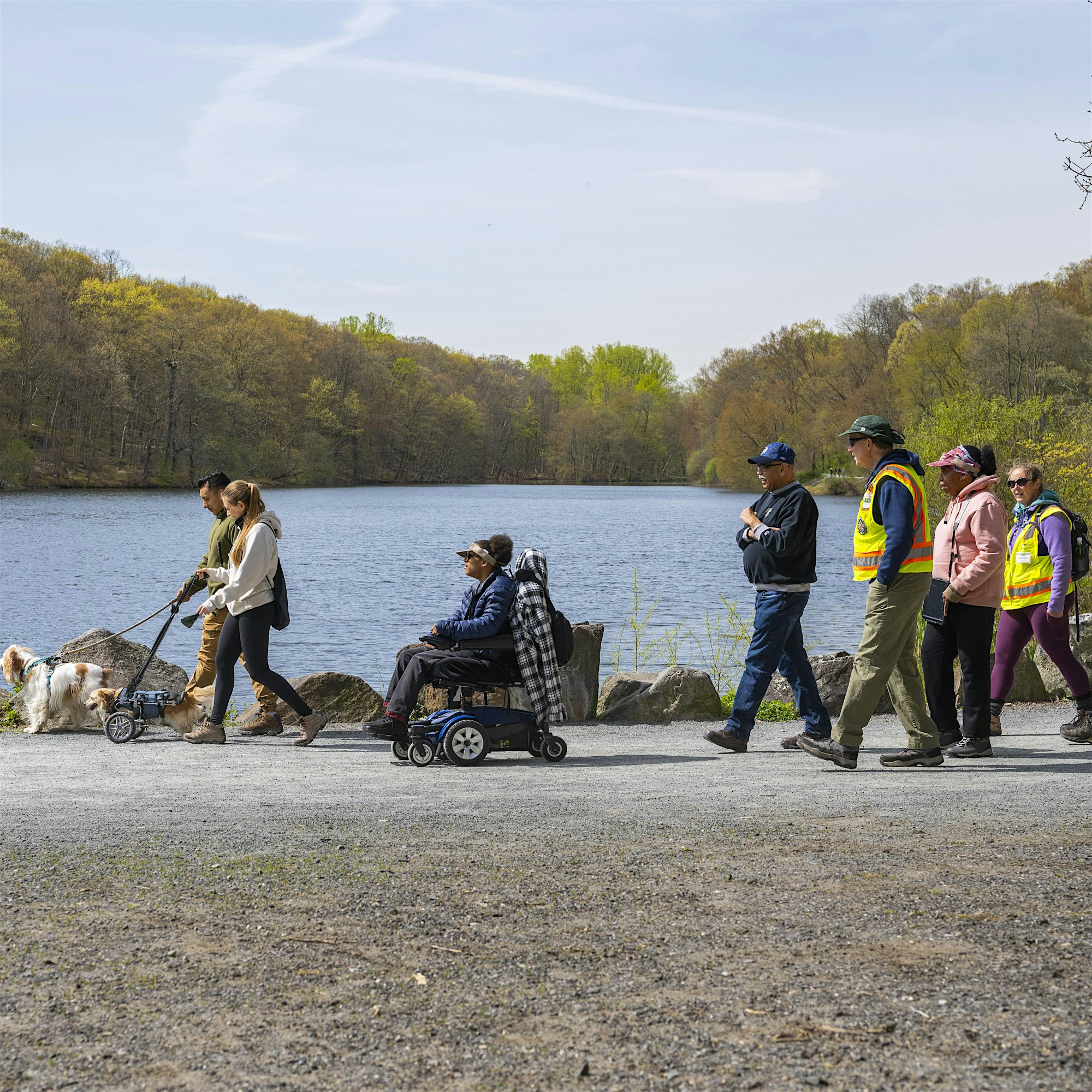 People walk along a lakefront path; some wear high-visibility vests, others in jackets, with a dog and a wheelchair user in view.