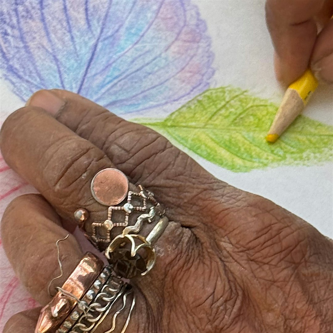 Close-up of a dark-skinned hand wearing several metallic rings while coloring with a yellow pencil on colorful paper.