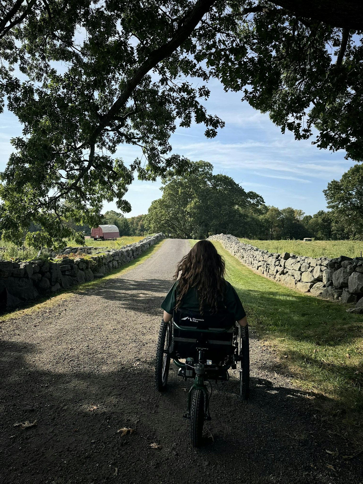 Person in a wheelchair on a gravel path between stone walls, with trees overhead and a distant red barn under a blue sky.