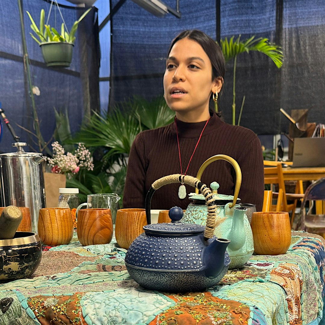 Woman with dark hair in a burgundy turtleneck sits at a table with blue teapot and wooden cups, in a cozy plant-filled setting describing a tea demonstration.