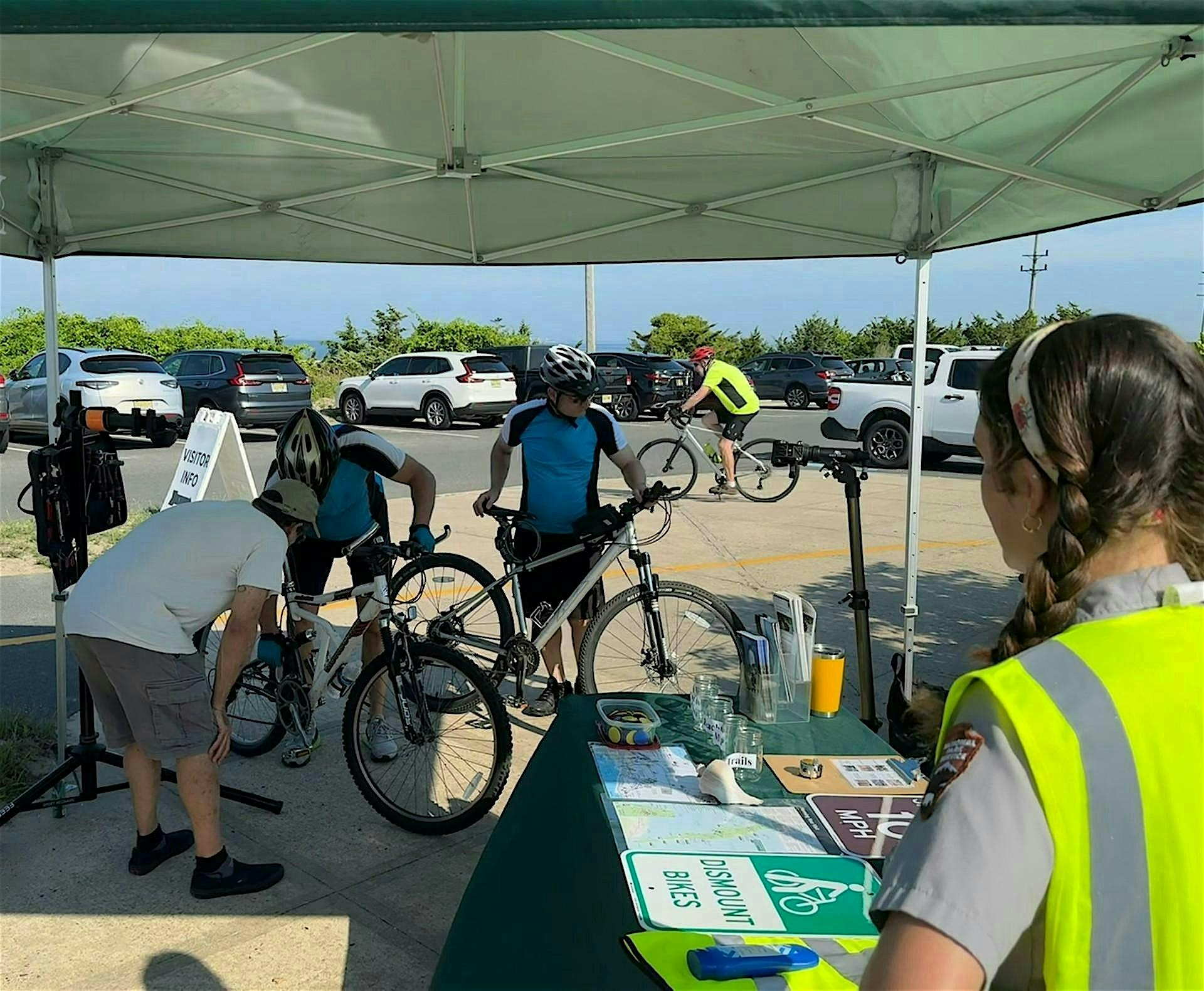 Cyclists under a canopy at an outdoor bike event with maps on a table and volunteers wearing high-visibility vests.