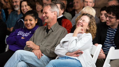 Audience members seated and smiling at a live event; foreground shows a man in a gray shirt laughing with two women, one in a purple hoodie.