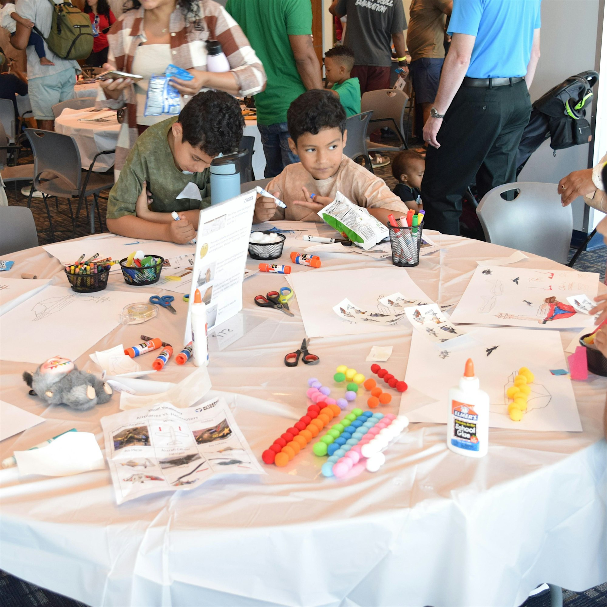 Two young boys sit at a round craft table coloring and cutting paper with markers, glue, and scissors while others stand around in a busy event space.