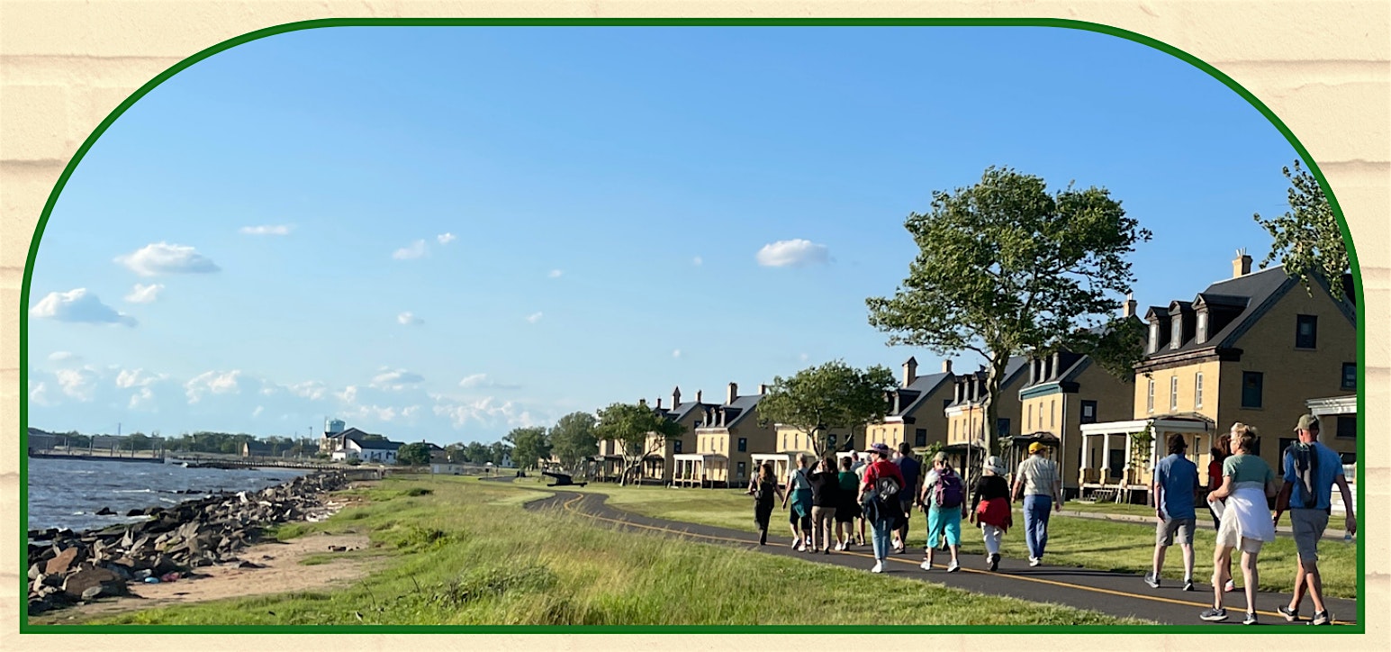 Group of people walking along a seaside promenade with houses and trees on a sunny day by the water's edge.