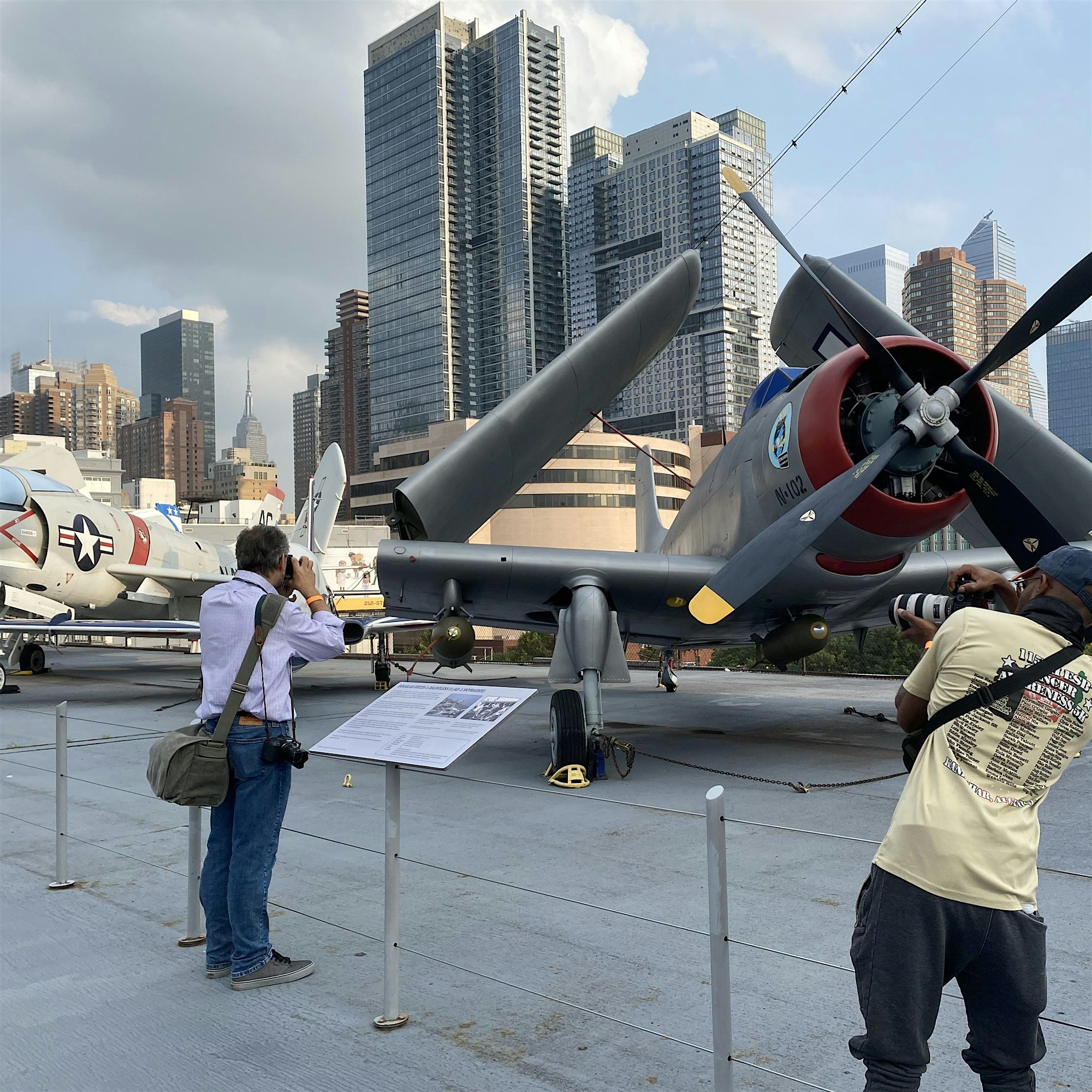 Photographer with camera beside a vintage propeller plane on the tarmac, city skyscrapers in the background.