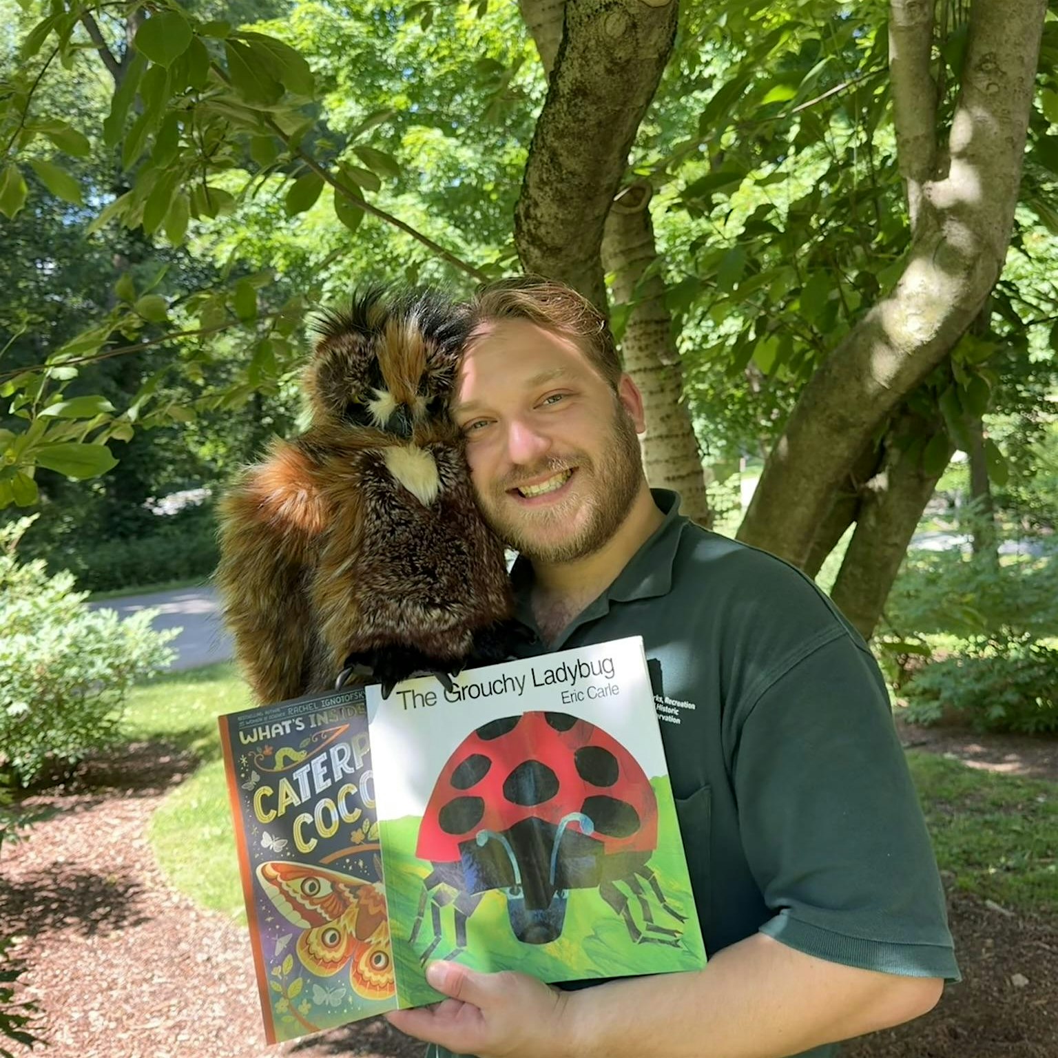 Smiling man outdoors with a large fluffy owl perched on his shoulder, holding colorful children's books (The Grouchy Ladybug and other titles).