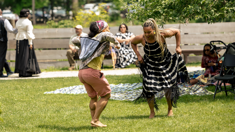 Two people dance barefoot on a grassy park, surrounded by onlookers and a blanket on the ground during an outdoor gathering.
