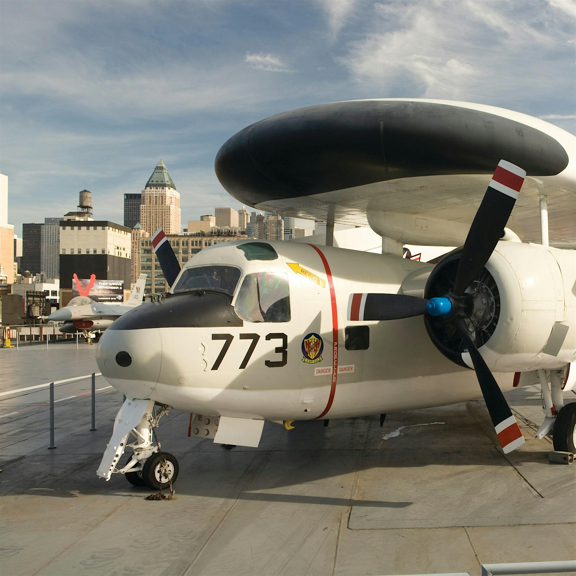 Close-up of a large aircraft nose and rotor blades on a tarmac with city skyscrapers in the background.