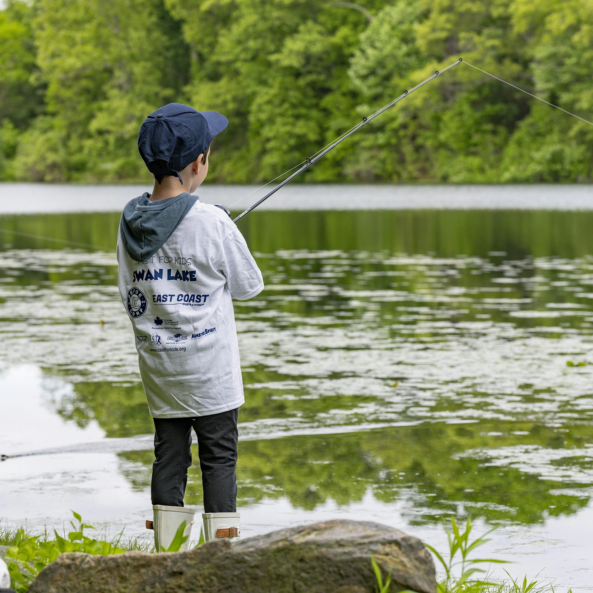 Back view of a child wearing a cap and waders fishing by a lake on a rocky shore with green vegetation in the background