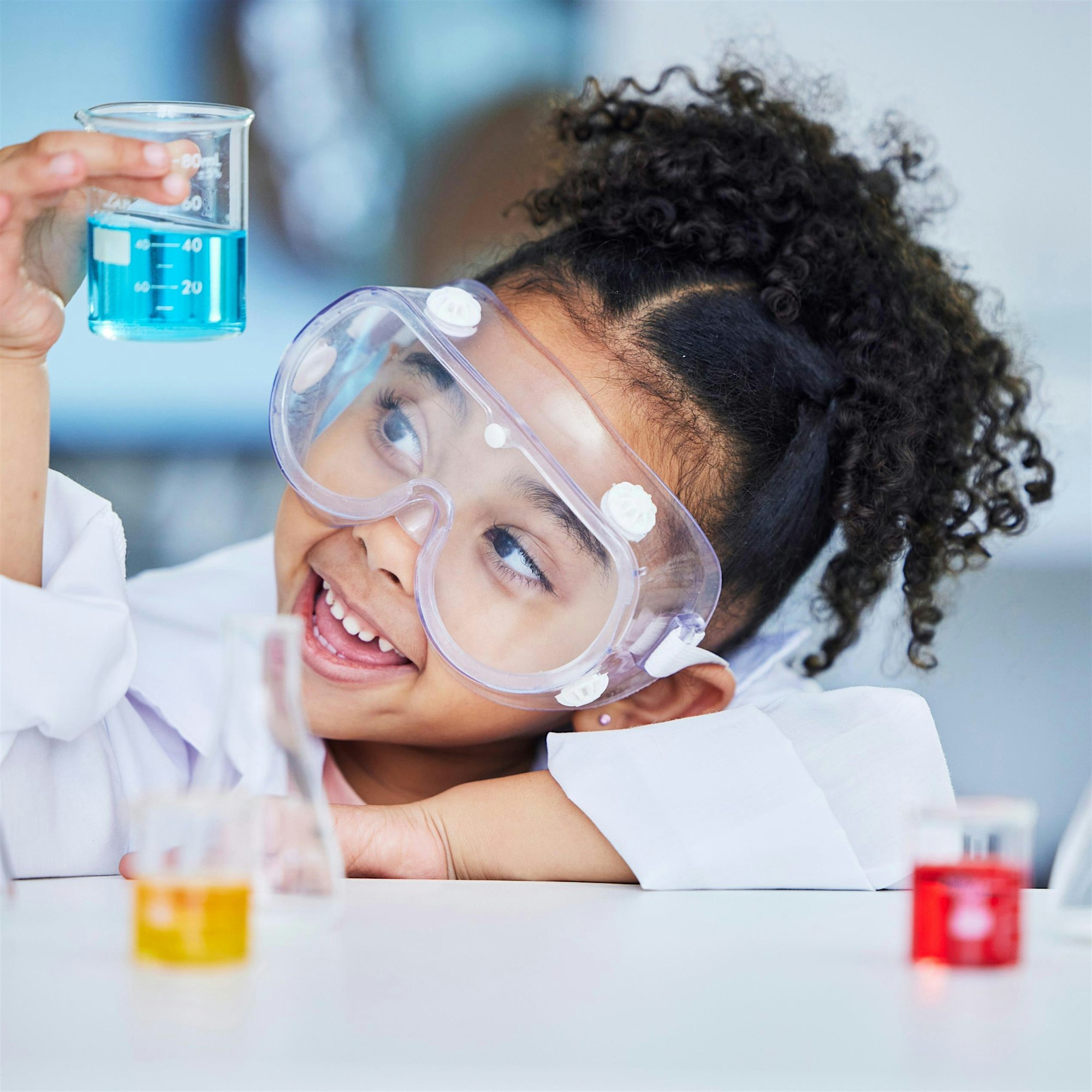 Young student in safety goggles holding a beaker with bright blue liquid in a lab environment.
