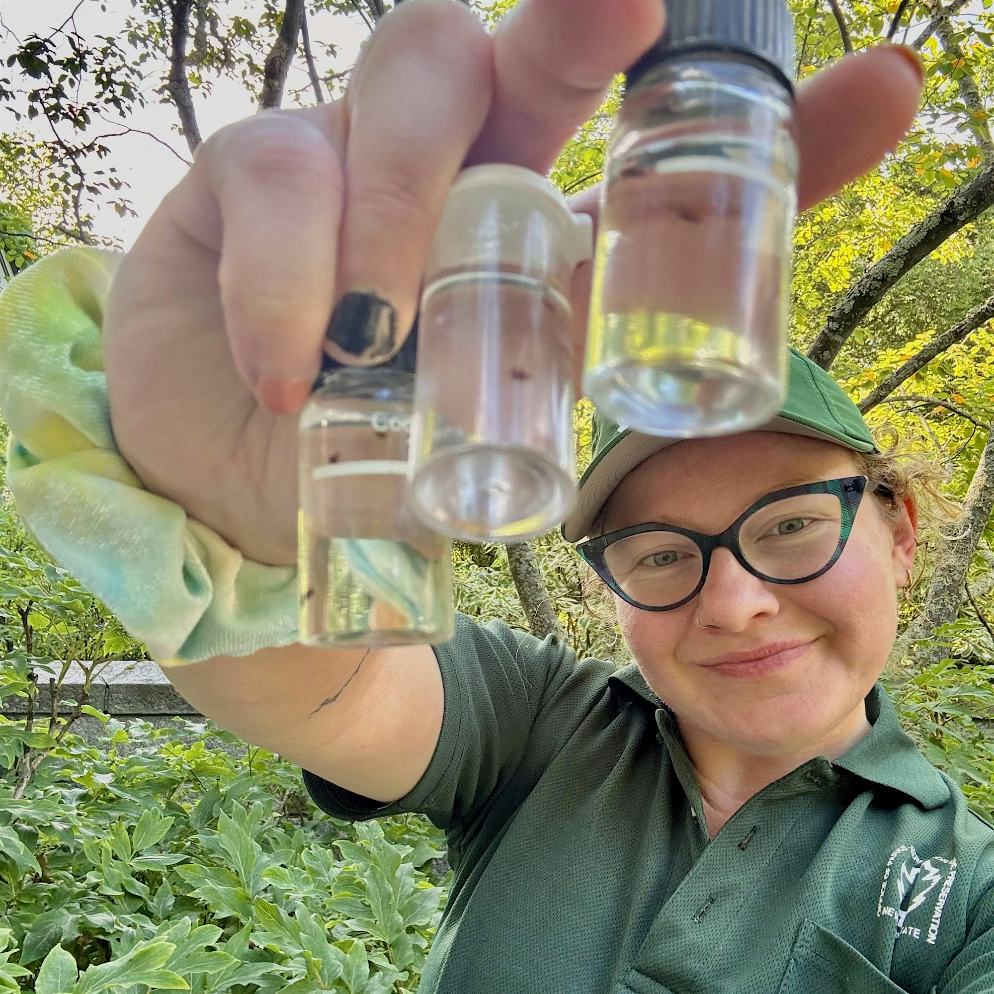 Person in a green shirt raises two small glass vials outdoors, with leafy trees in the background.