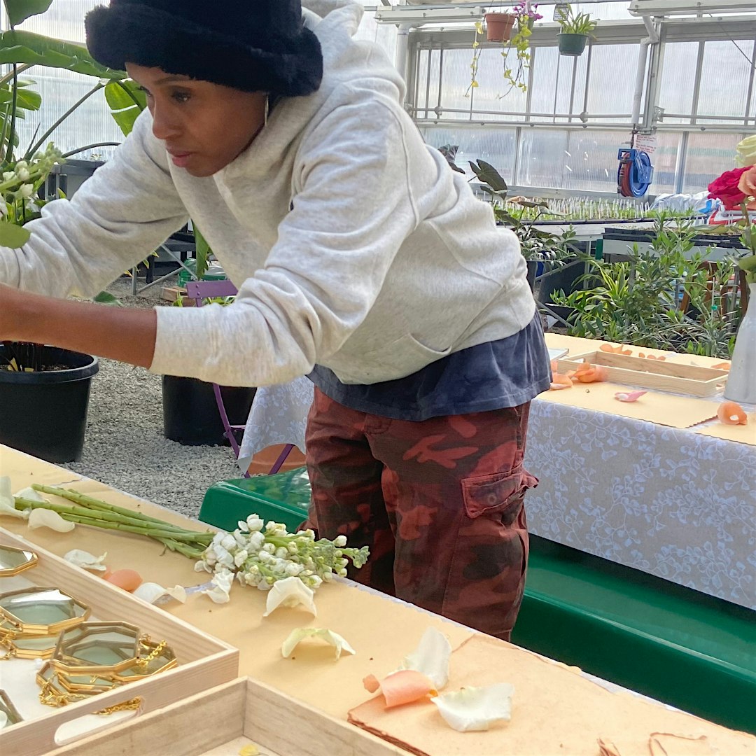 Person in a gray hoodie and black hat leans over a table in a greenhouse, arranging flowers and stems on a tray.