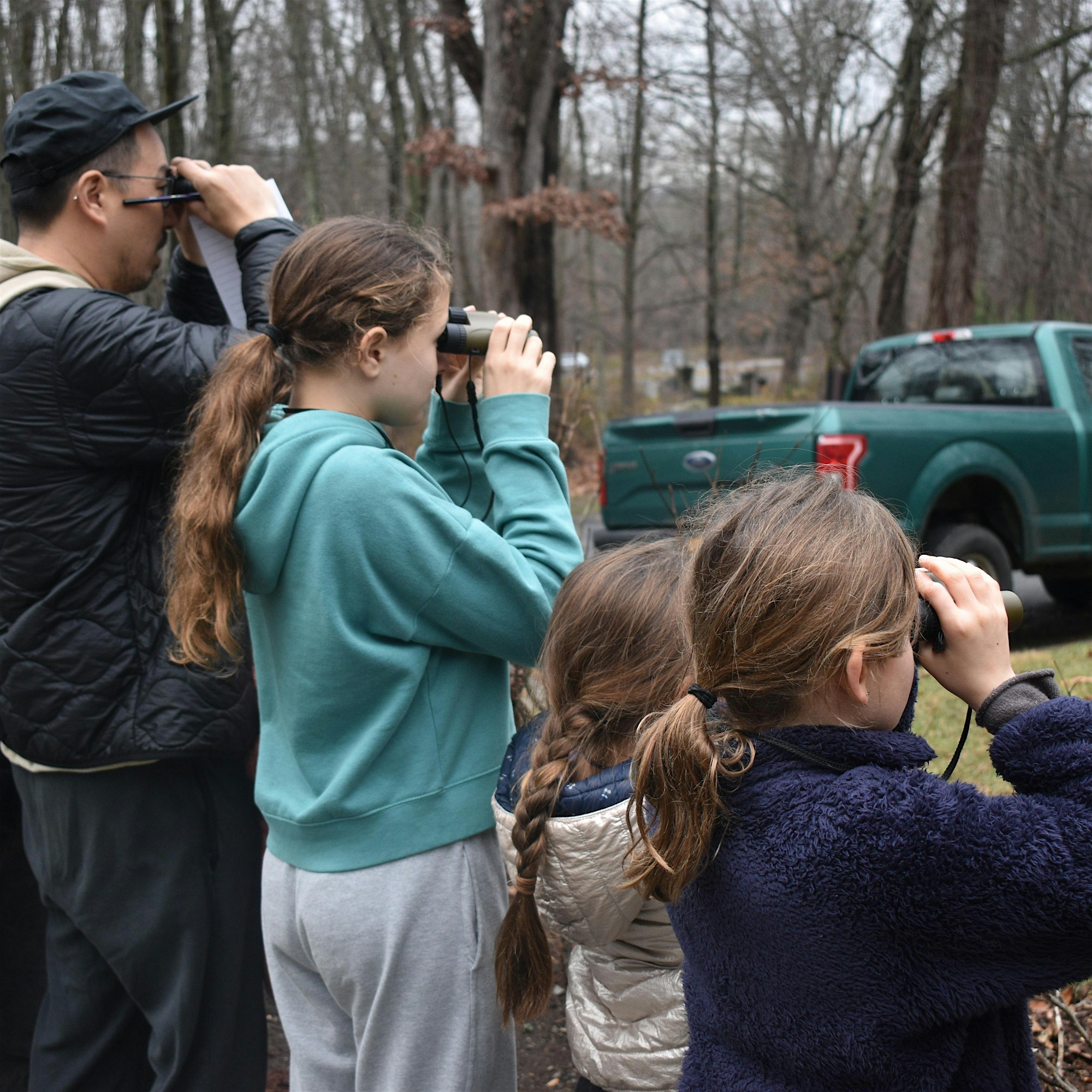 ee5060a0cff74c2254e06dda3873de7a - PARENTGUIDENEWS Children outdoors in a wooded area; one girl with binoculars and braided hair watches intently while others look on, a man holds paperwork nearby and a green truck is in the background.