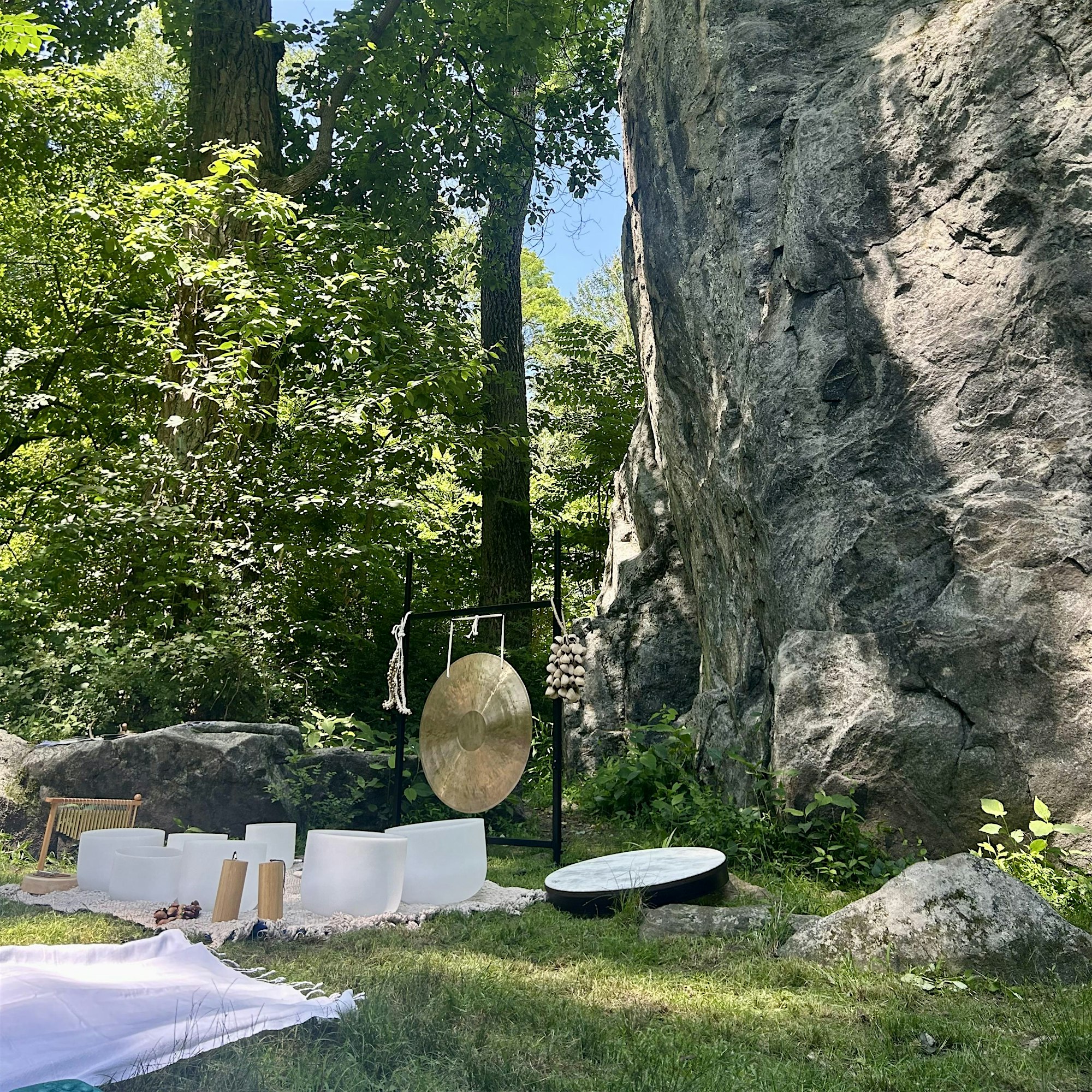 Outdoor seating area set up against a rocky cliff with a large gong and hanging garlic bulbs, surrounded by trees and greenery.