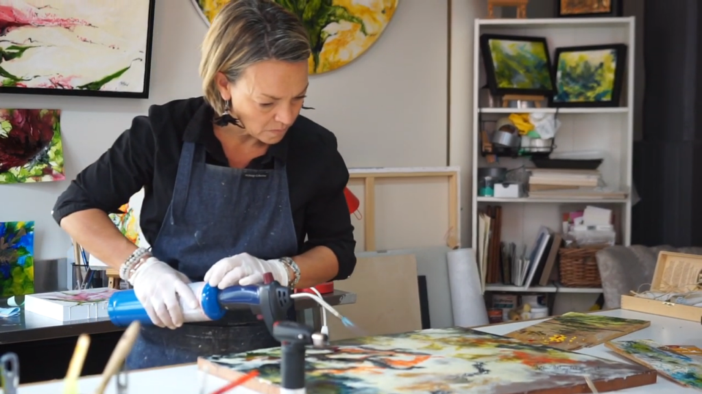 Female artist wearing a dark apron and gloves, using a blue rotary tool to work on a colorful painting in her studio.