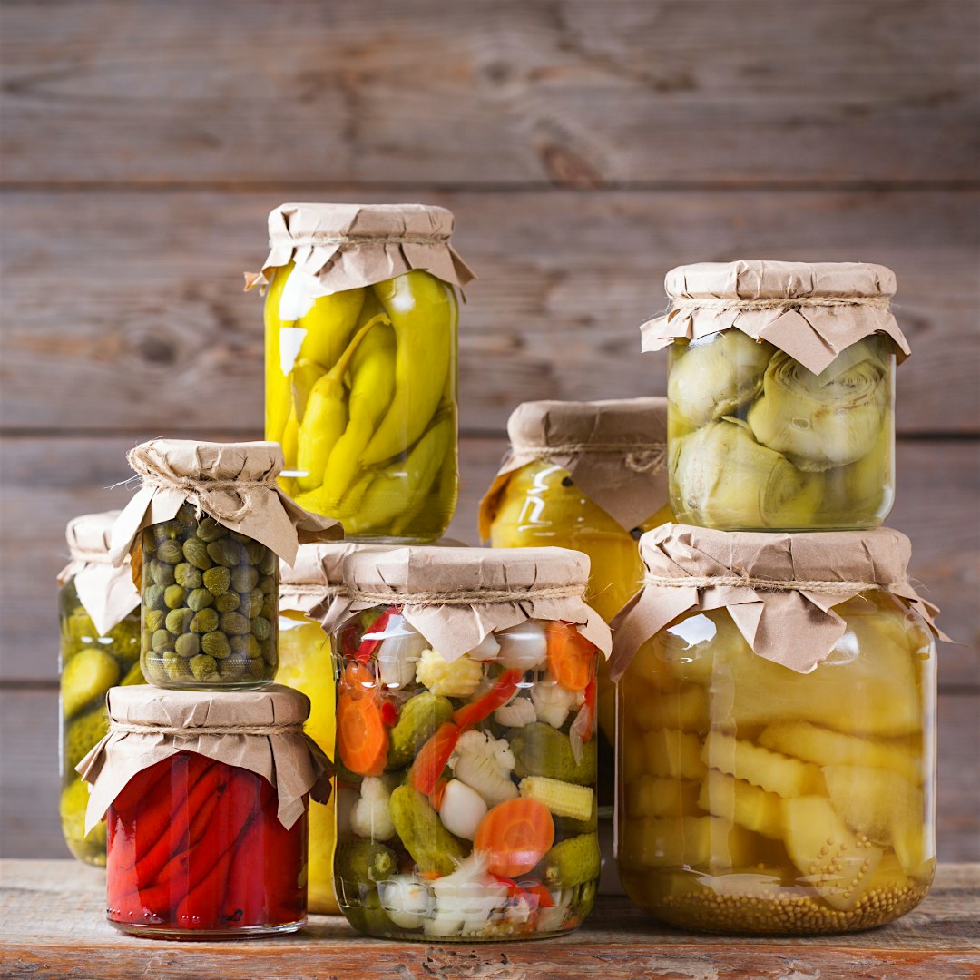 Assortment of glass jars filled with pickled vegetables (peppers, cucumbers, onions) sealed with paper lids on a wooden table.