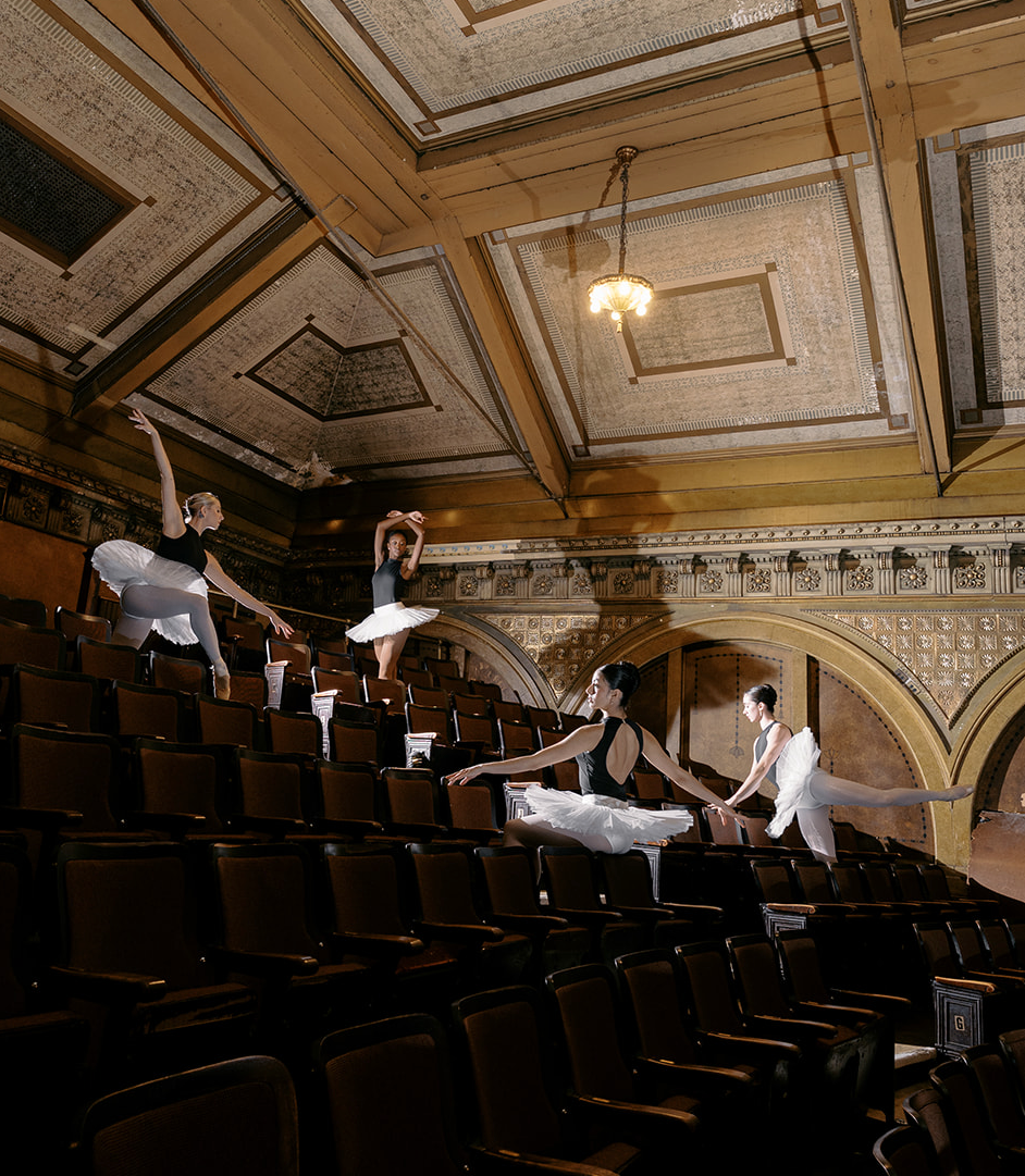 Four ballerinas in black leotards and white tutus dance on empty theater seats in a historic ornate auditorium.
